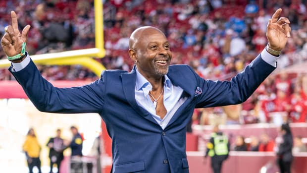 Former 49ers receiver Jerry Rice, wearing a suit, holds his hands up on the field before a game